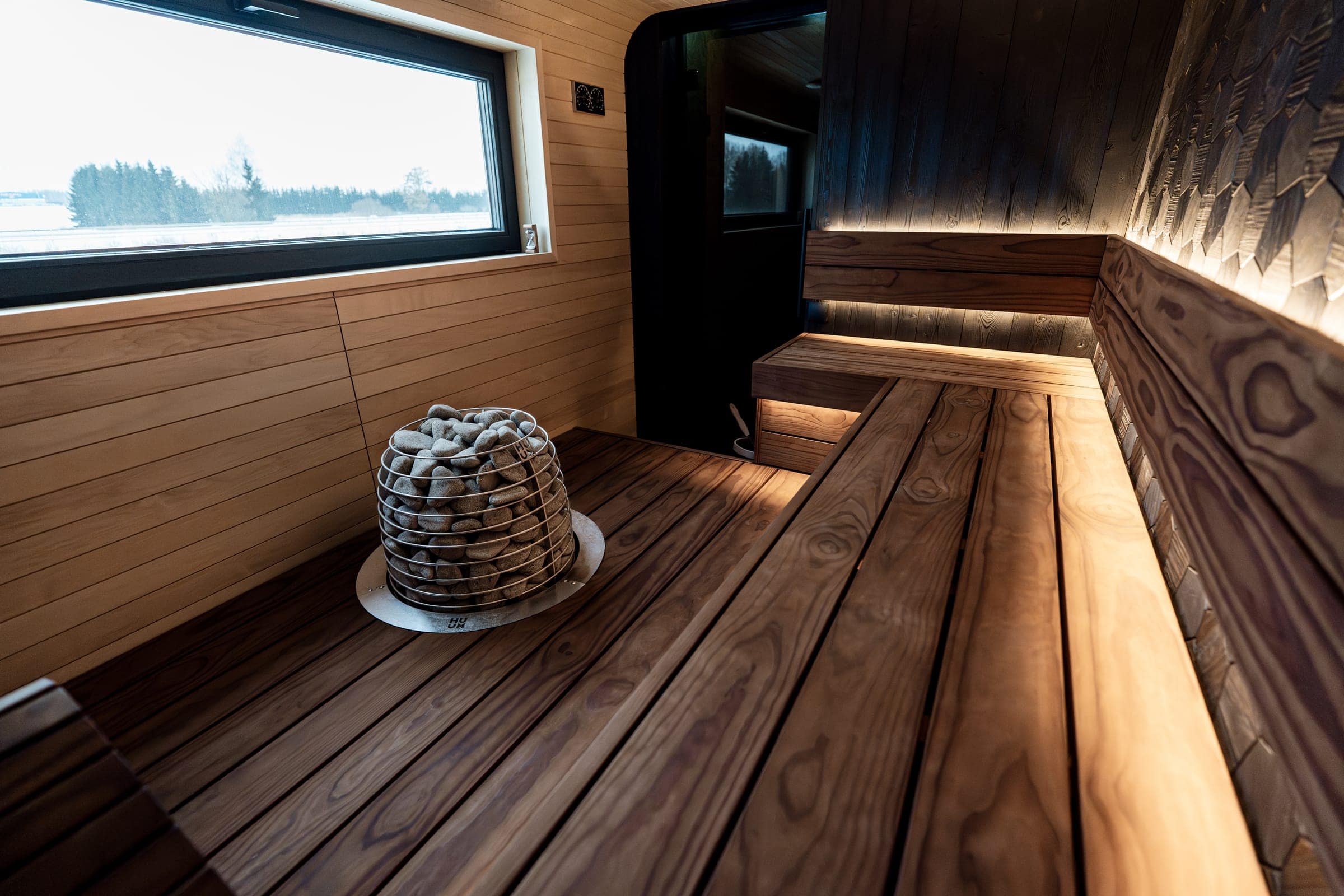 HUUM HIVE set into the floor of a wood sauna with lit benches and a window onto snow