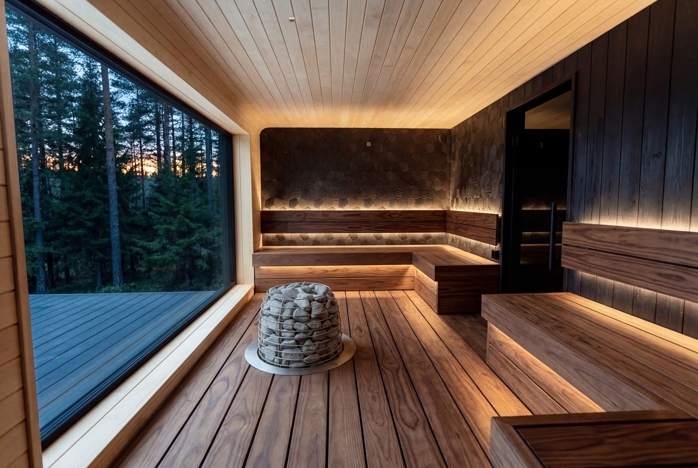 HUUM HIVE heater in the middle of a long dark-wood sauna with a floor-to-ceiling window onto a forest at dusk