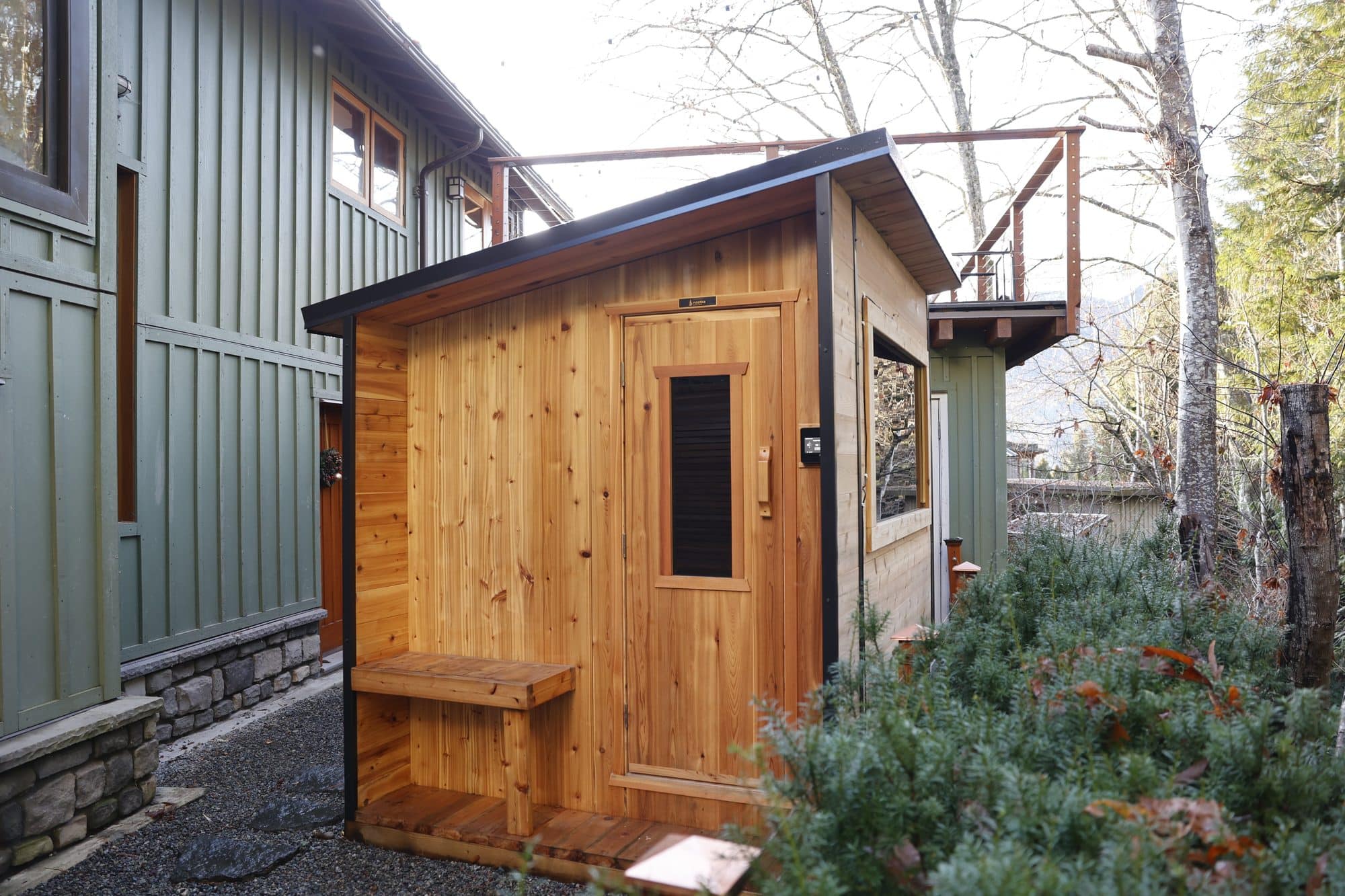 Nootka Cube sauna with a sloped roof and an outdoor bench beside a green-sided house