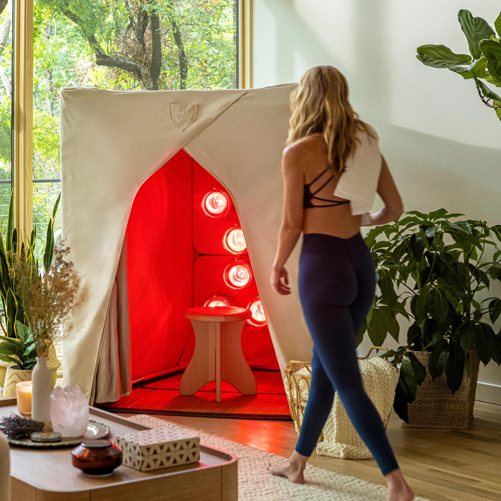 A woman with a towel walking toward a glowing FireLight sauna in a room full of plants