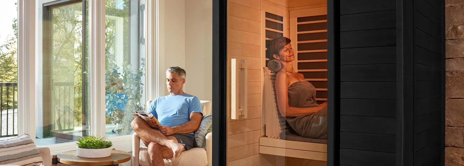 A woman wrapped in a towel leans back inside a black Sun Home Solstice behind its glass door while a man reads in an armchair by the window