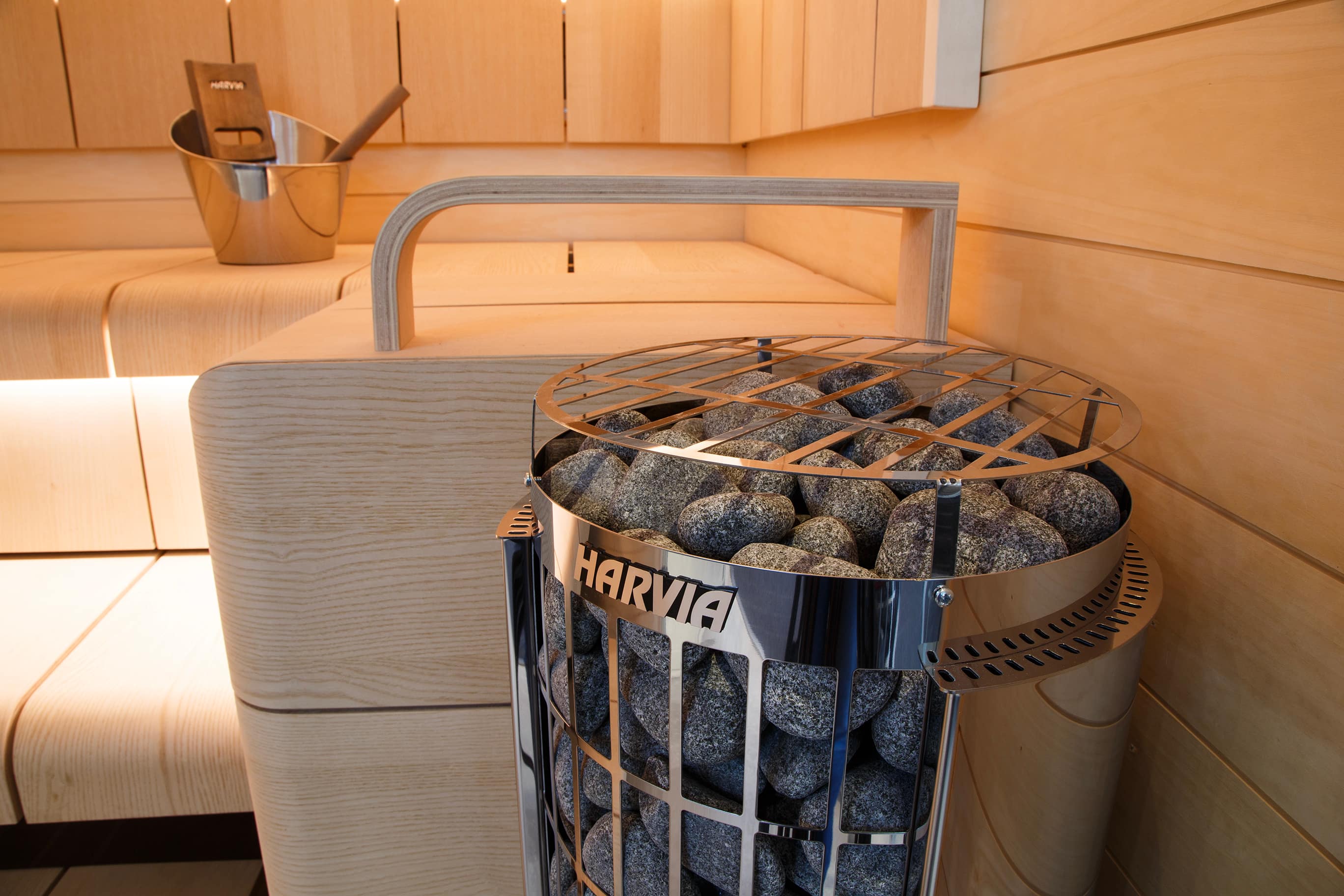 Harvia Cilindro Half heater behind a curved bench rail in a pale wood sauna with a steel bucket on the bench