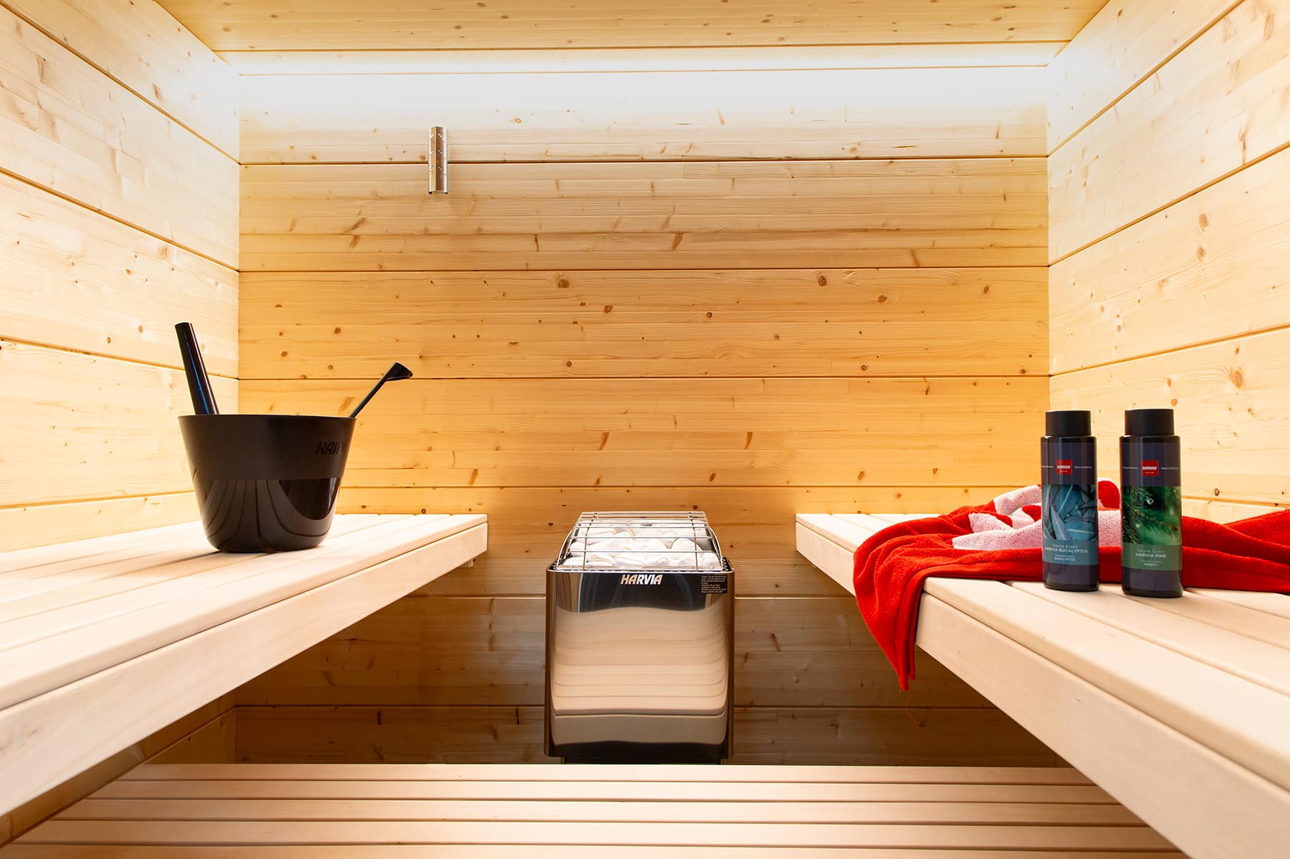 Harvia KIP W stainless heater between two spruce benches holding a bucket and bottles in a lit sauna