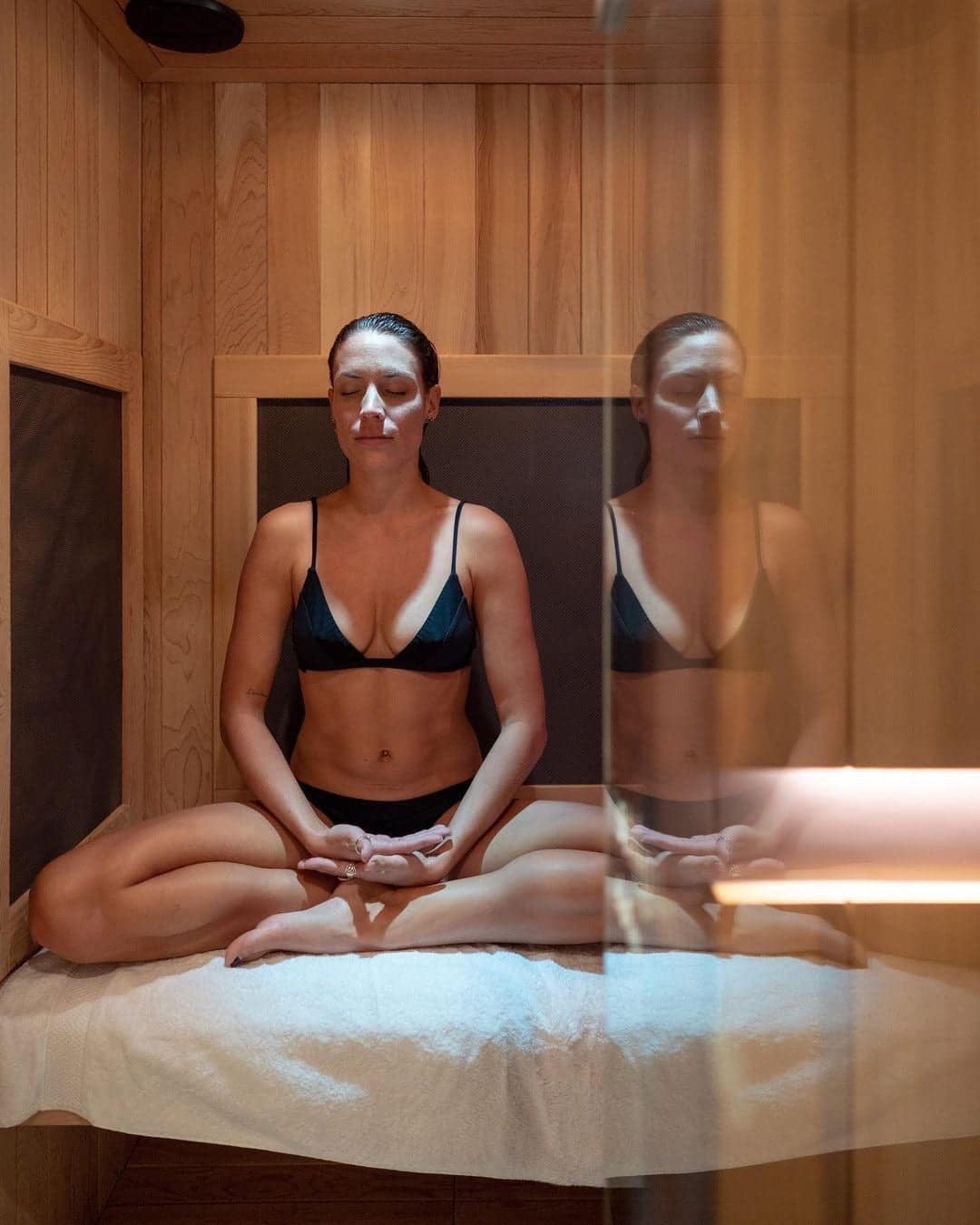 A woman meditating cross-legged inside a Sunlighten mPulse Conquer with her reflection in the glass