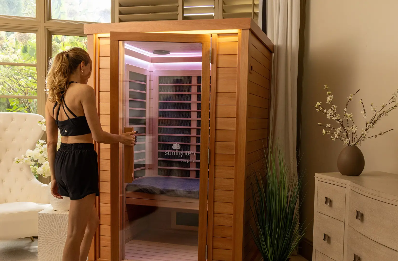 A woman opening the glass door of a Sunlighten Amplify II cabin in a sunlit room